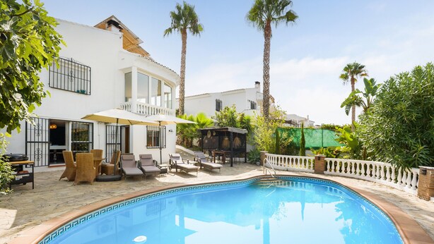 A swimming pool with lounge chairs and umbrellas, surrounded by palm trees and a white building with balconies. - Holiday Home 'Serena Vista' with Mountain View, Wi-Fi, and Air Conditioning (Alcaucín)