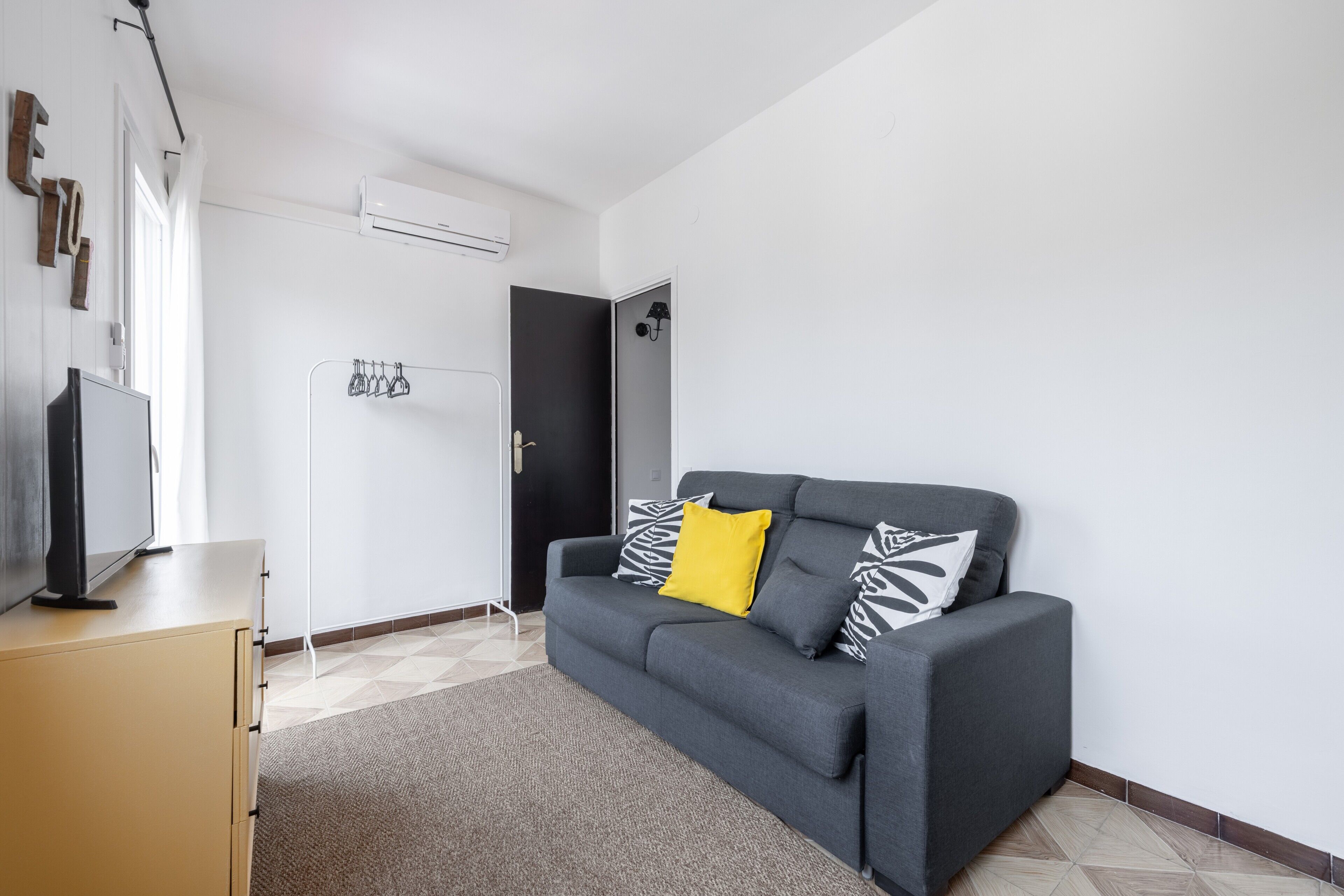 Un saló modern amb un sofà gris, una televisió de pantalla plana sobre una estanteria, un penjador i una porta que condueix a una altra habitació.