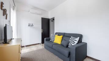 Un saló modern amb un sofà gris, una televisió de pantalla plana sobre una estanteria, un penjador i una porta que condueix a una altra habitació.