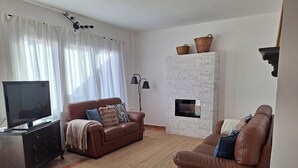Un salón con un sofá de cuero marrón, un televisor sobre un mueble, una chimenea y un amplio ventanal con cortinas.