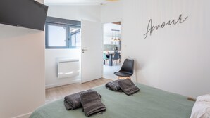 A modern bedroom with a green bed, a wall-mounted TV, a ceiling light, and a view into the kitchen.
