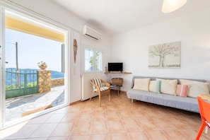 A living room with a sofa, a chair, a television, a wall-mounted artwork, a window, and a door leading to a balcony.