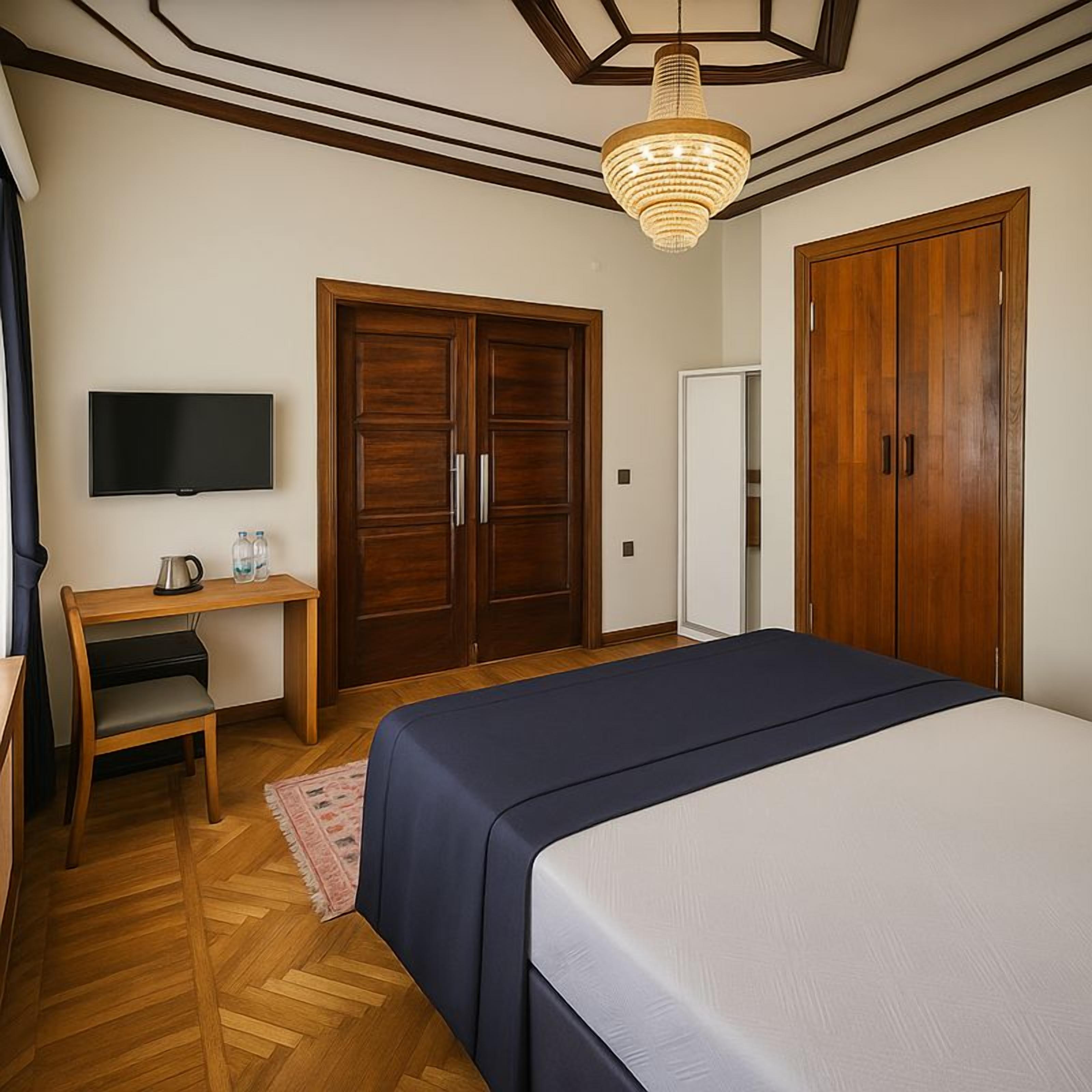 A bedroom with a bed, wooden wardrobe, a desk with a chair, a television, and a hanging light fixture.