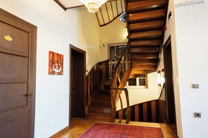 Escada de madeira com corrimão, porta com placa de número e tapete vermelho estampado no chão.
