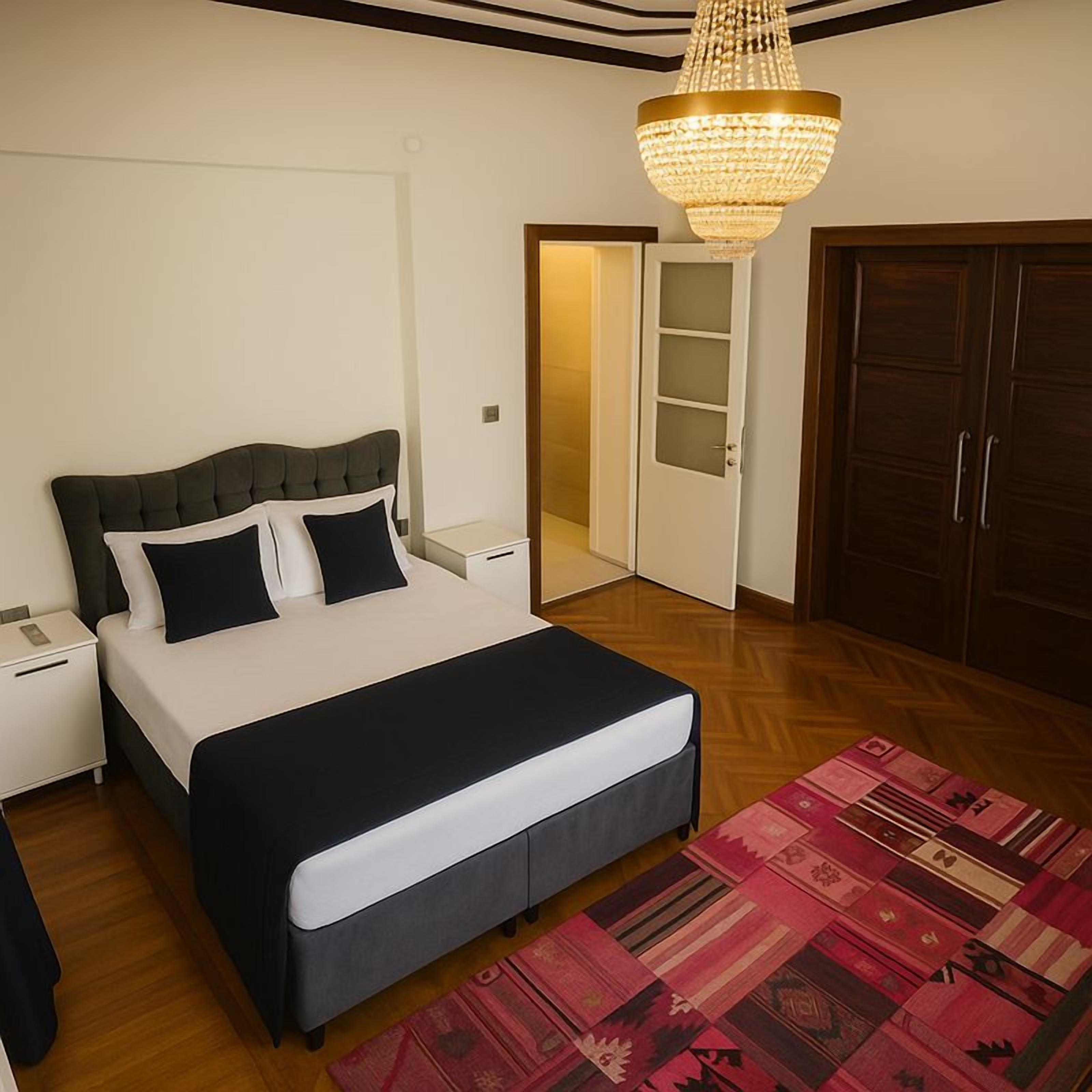 A bedroom with a bed, nightstands, a chandelier, a wardrobe, and a door leading to another room.