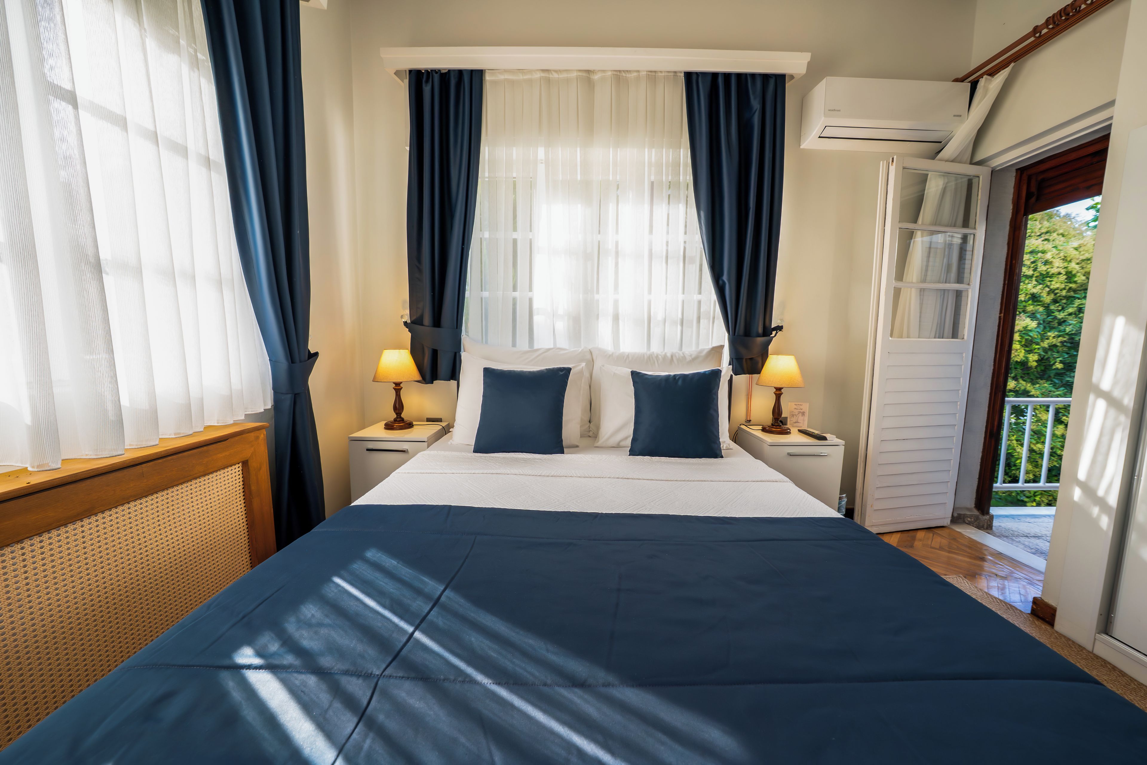 Habitación de hotel con cama, dos mesitas de noche, ventana con cortinas, equipo de aire acondicionado y puerta que da a un espacio exterior.
