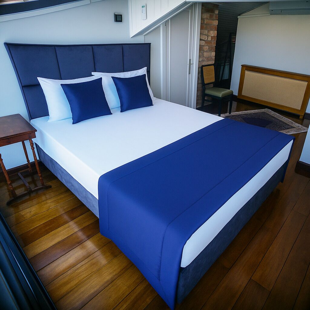 A neatly made bed with a blue and white bedspread, a wooden nightstand, and a chair in a room with wooden flooring and a brick wall.