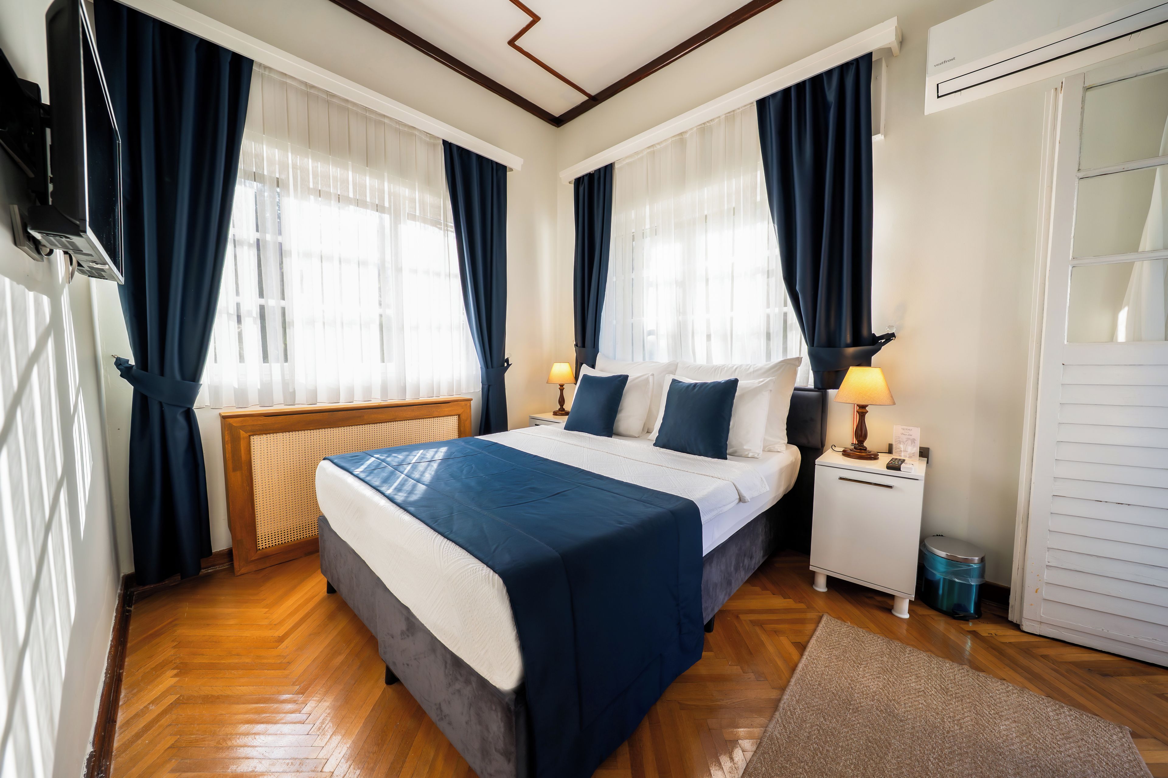 Una habitación de hotel con cama, mesitas de noche, lámpara, televisor y ventana con cortinas.