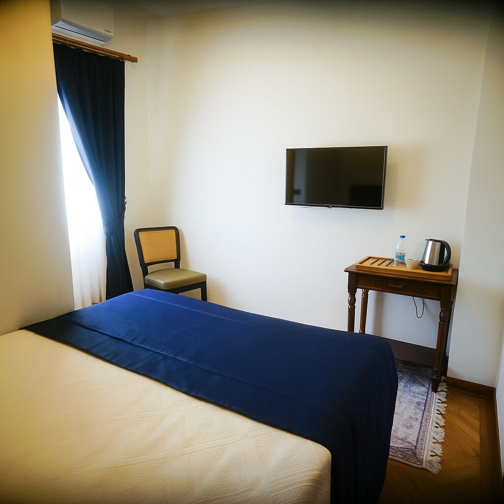 A hotel room with a bed, a chair, a TV, and a small table with a kettle and a bottle.