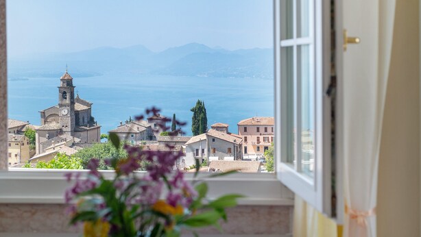 A view through a window with a clock tower, buildings, and a body of water. - Room 'Gemme Suite Bb' with Lake View, Balcony and Wi-Fi (Torri del Benaco)