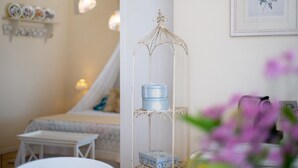 A room with a white bed, a small table, a decorative metal stand with boxes, and a framed botanical print on the wall. - Room 'Gemme Suite Bb' with Lake View, Balcony and Wi-Fi (Torri del Benaco)