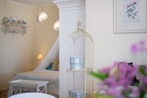 A room with a white bed, a small table, a decorative metal stand with boxes, and a framed botanical print on the wall.