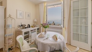 A dining area with a table set for two, a white chair, a small cabinet, and a view of the sea through a window. - Room 'Gemme Suite Bb' with Lake View, Balcony and Wi-Fi (Torri del Benaco)