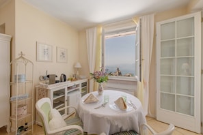 A dining area with a table set for two, a white chair, a small cabinet, and a view of the sea through a window.