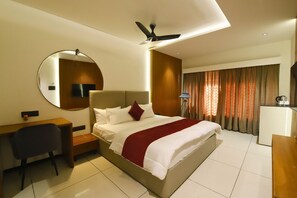 A hotel room with a large bed, a desk, a chair, a mirror, a ceiling fan, and a window with curtains. - Hotel Sanatana (Ujjain)