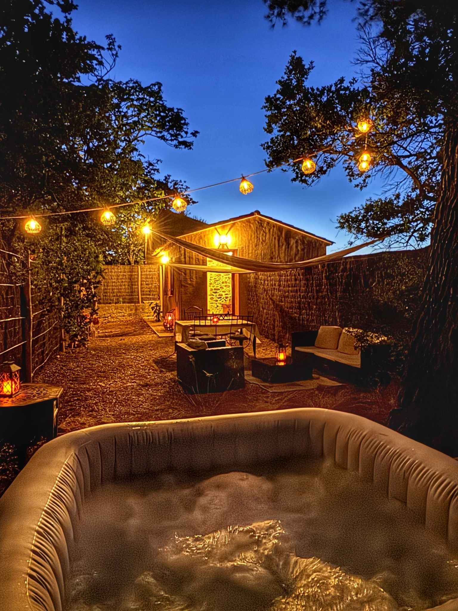 A hot tub in a backyard with a wooden house, string lights, and a stone wall.