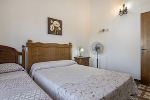 Un dormitorio con dos camas, cabeceros de madera, una mesita de noche con lámpara, un ventilador de pared y una puerta.