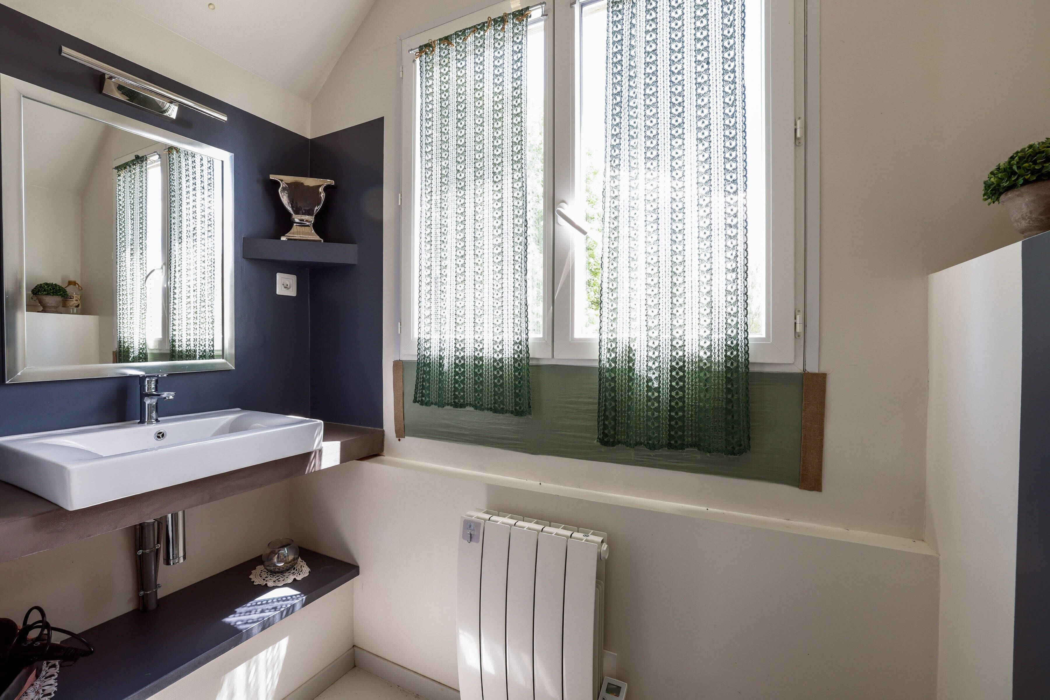 A bathroom with a white sink, a mirror, a radiator, and a window with patterned curtains.