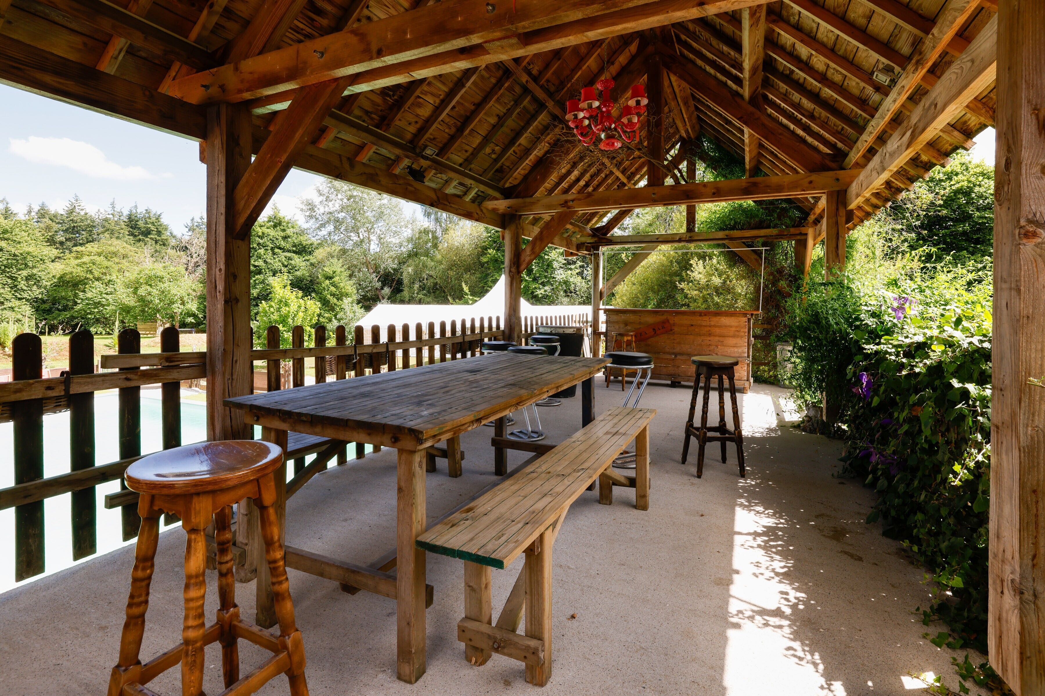 A wooden pavilion with a table and benches, surrounded by a wooden fence and greenery.