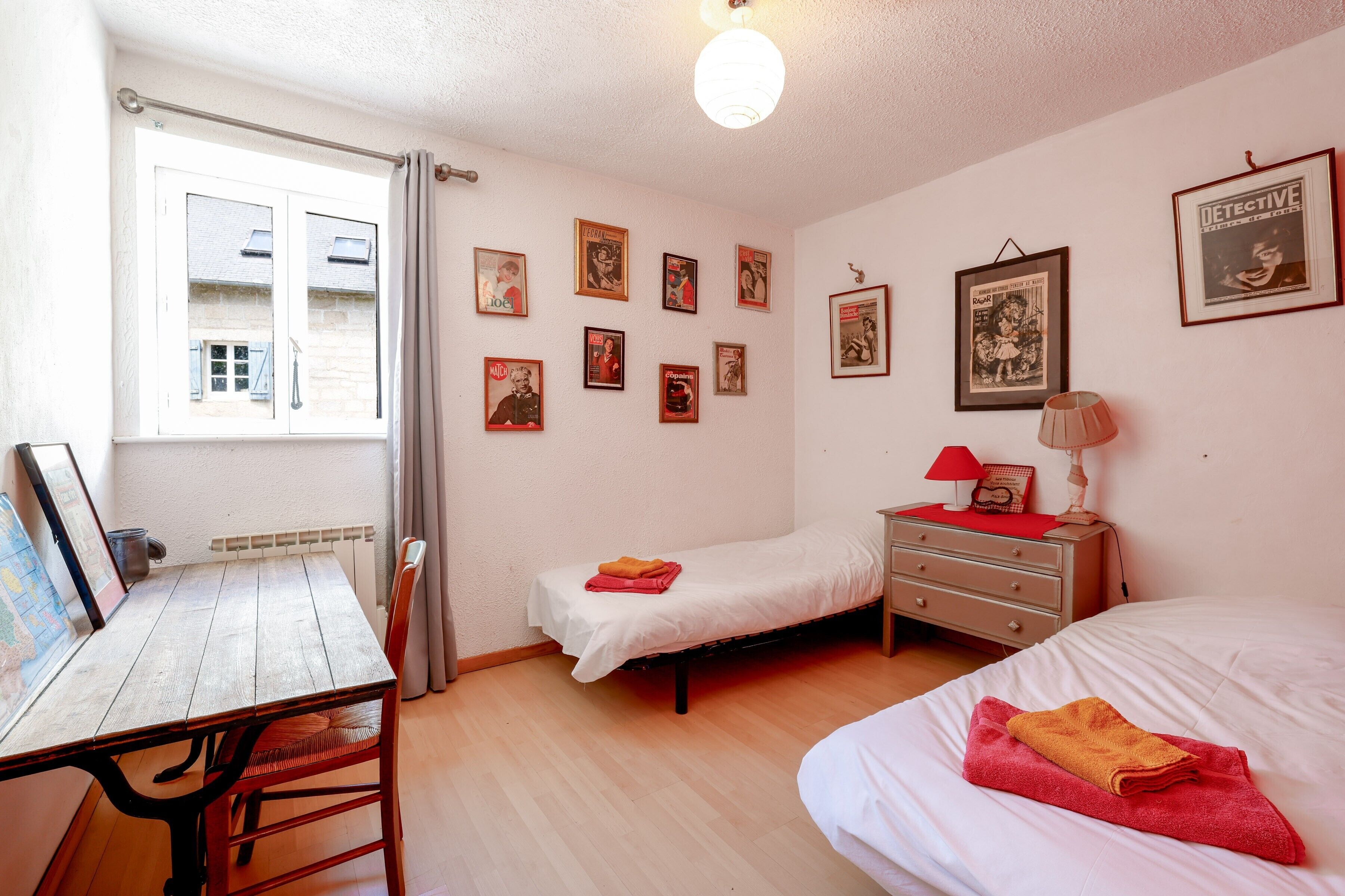Een kamer met een houten tafel, een bed, een klein nachtkastje met een lamp en een raam met gordijnen.