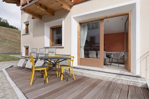 Una terraza de madera con una mesa y sillas, una puerta corrediza de vidrio que da a un comedor interior, y una ladera en el fondo.
