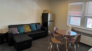 Un espace de vie compact comprenant un canapé, une petite table et des chaises. Un réfrigérateur est placé contre le mur, et il y a une fenêtre avec des stores.