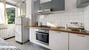 Une cuisine compacte avec des meubles blancs, un four encastré, un évier en acier inoxydable et une fenêtre avec des rideaux.