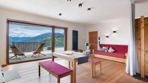 Un interior moderno con suelo de madera, un sofá rojo, una mesa de comedor y vistas a montañas a través de grandes ventanales.