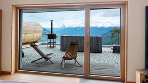 Una habitación con un amplio ventanal corredera que da a una terraza exterior con una silla de madera, una mesa y vistas a montañas nevadas.