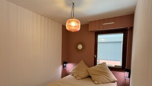 A bedroom with a bed, a hanging light fixture, a window with blinds, and a wall-mounted decorative item. - T2bis - Cap d'Agde - 40 m² with terrace, swimming pool, parking & 200 m from the beach (Agde)