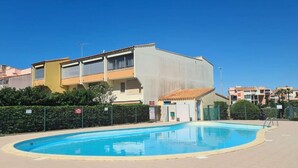 A swimming pool surrounded by buildings with balconies, a clear blue sky, and a fenced area. - T2bis - Cap d'Agde - 40 m² with terrace, swimming pool, parking & 200 m from the beach (Agde)