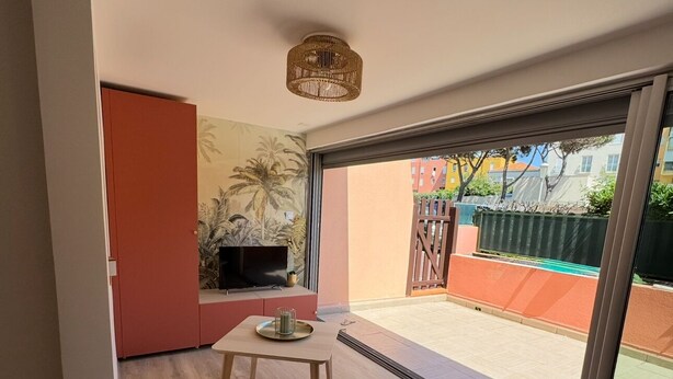 A modern living room with a sofa, a small table, and a TV. There is a sliding glass door leading to an outdoor area with a view of buildings and a fence. - T2bis - Cap d'Agde - 40 m² with terrace, swimming pool, parking & 200 m from the beach (Agde)