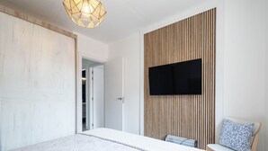 A bedroom with a bed, a TV mounted on a wooden panel wall, a chair with a cushion, and a door leading to another room.