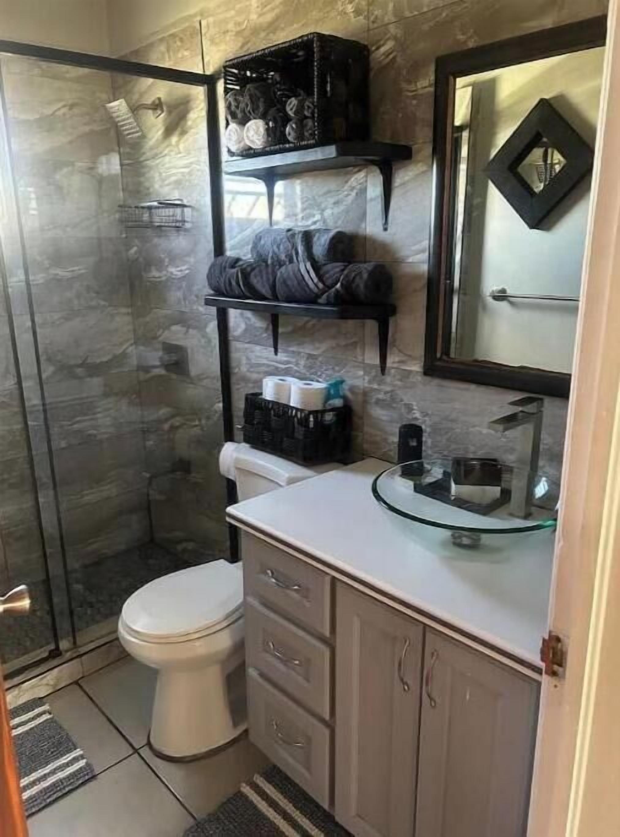 A bathroom with a glass shower enclosure, a toilet, a vanity with drawers, a mirror, and shelves with toiletries.