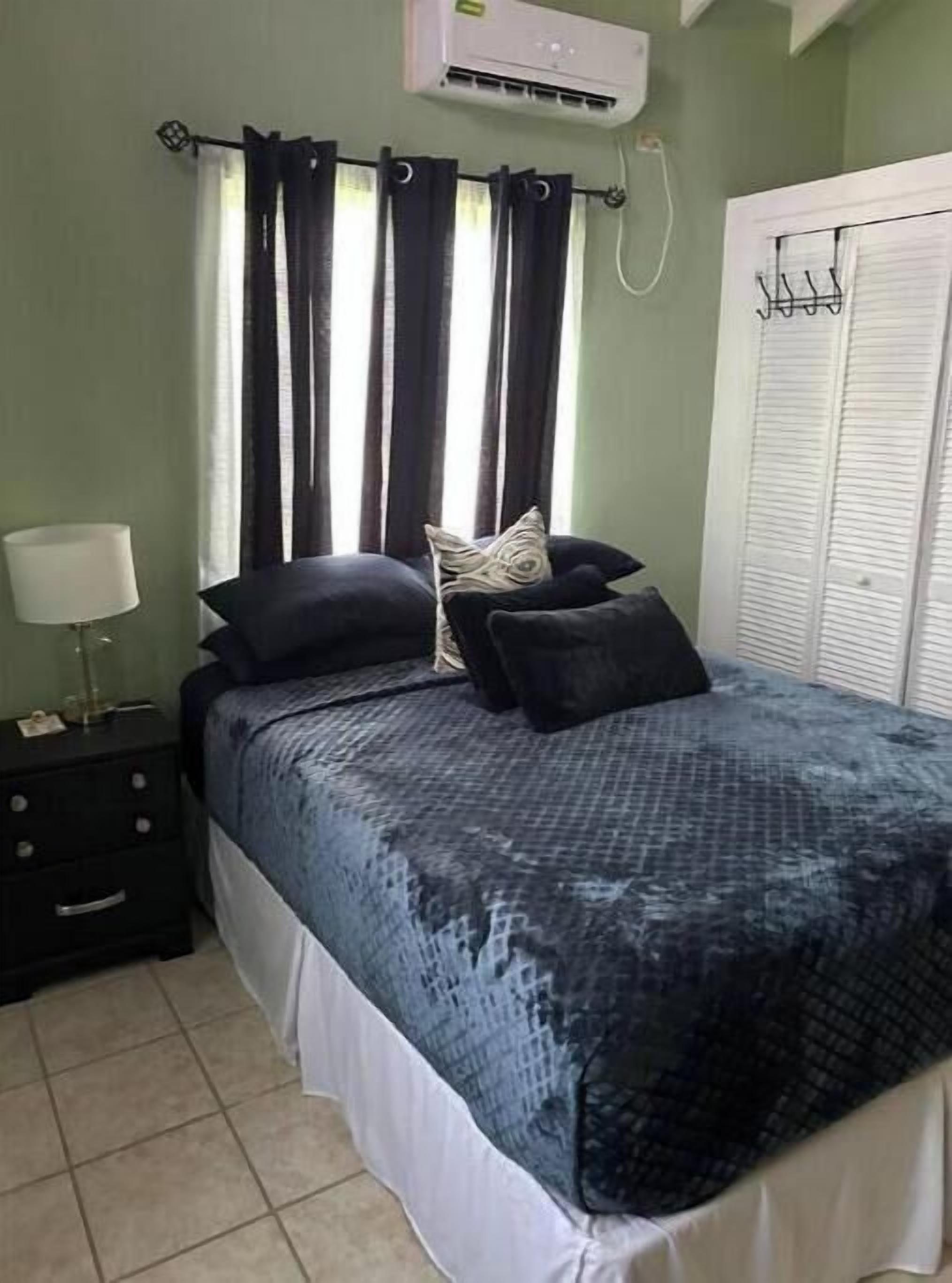 A bedroom with a bed, a nightstand, a lamp, a window with curtains, an air conditioning unit, and a closet.