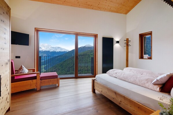 A room with a bed, a wooden bench, a TV, and a large sliding glass door leading to a mountain view.