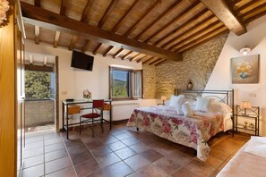 Un dormitorio con pared de piedra, vigas de madera en el techo, cama con ropa de cama floral, escritorio con silla y televisor montado en la pared.