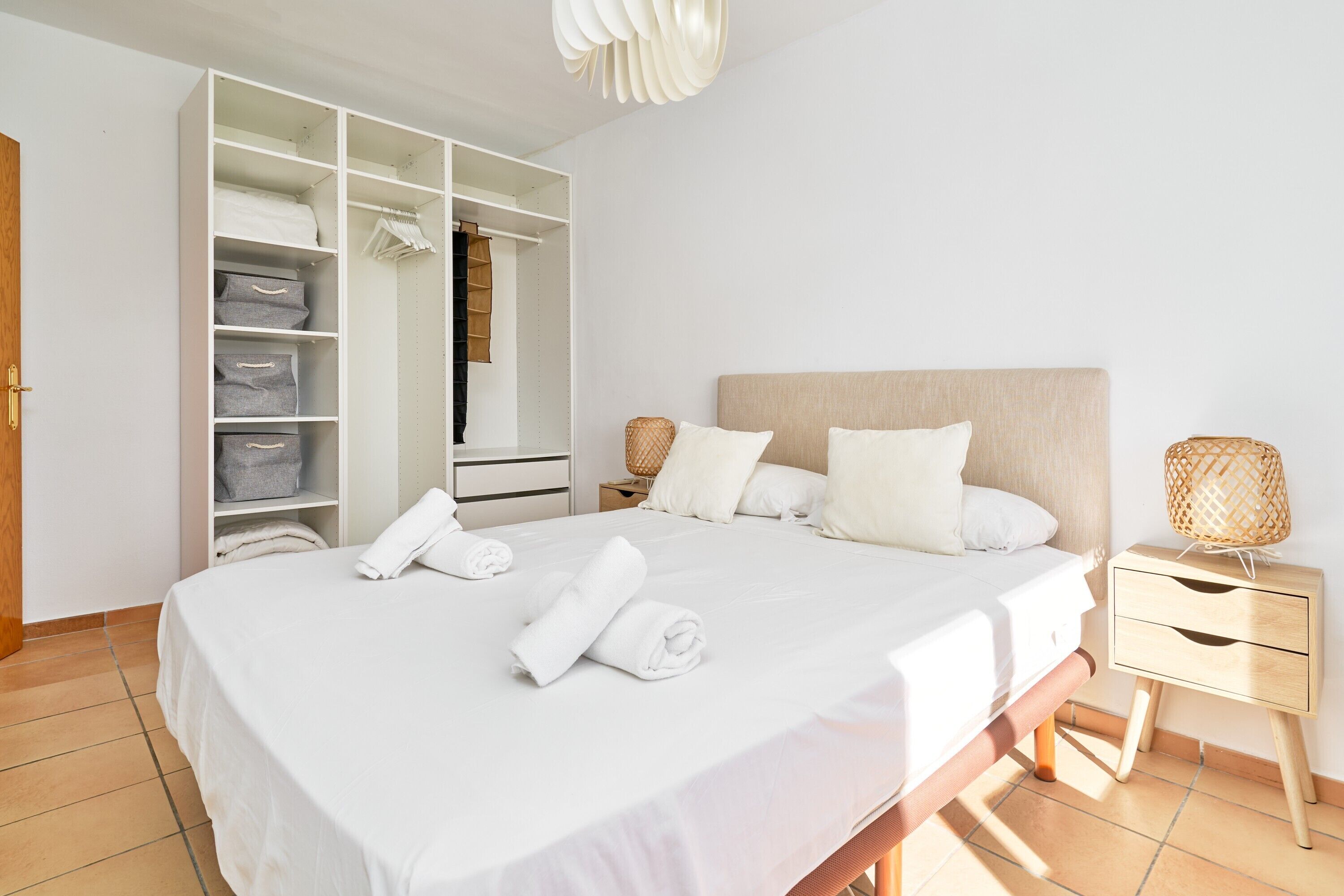 A neatly organized bedroom with a large bed, a wardrobe with multiple compartments, a bedside table with a lamp, and a nightstand.