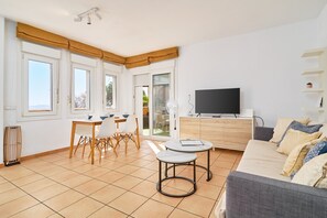 A modern living room with a sofa, a dining table with chairs, a TV on a wooden cabinet, and large windows with curtains.