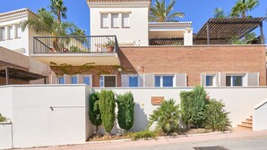 Un edificio de dos plantas con un balcón, fachada de ladrillo y una pared blanca con un pequeño jardín.
