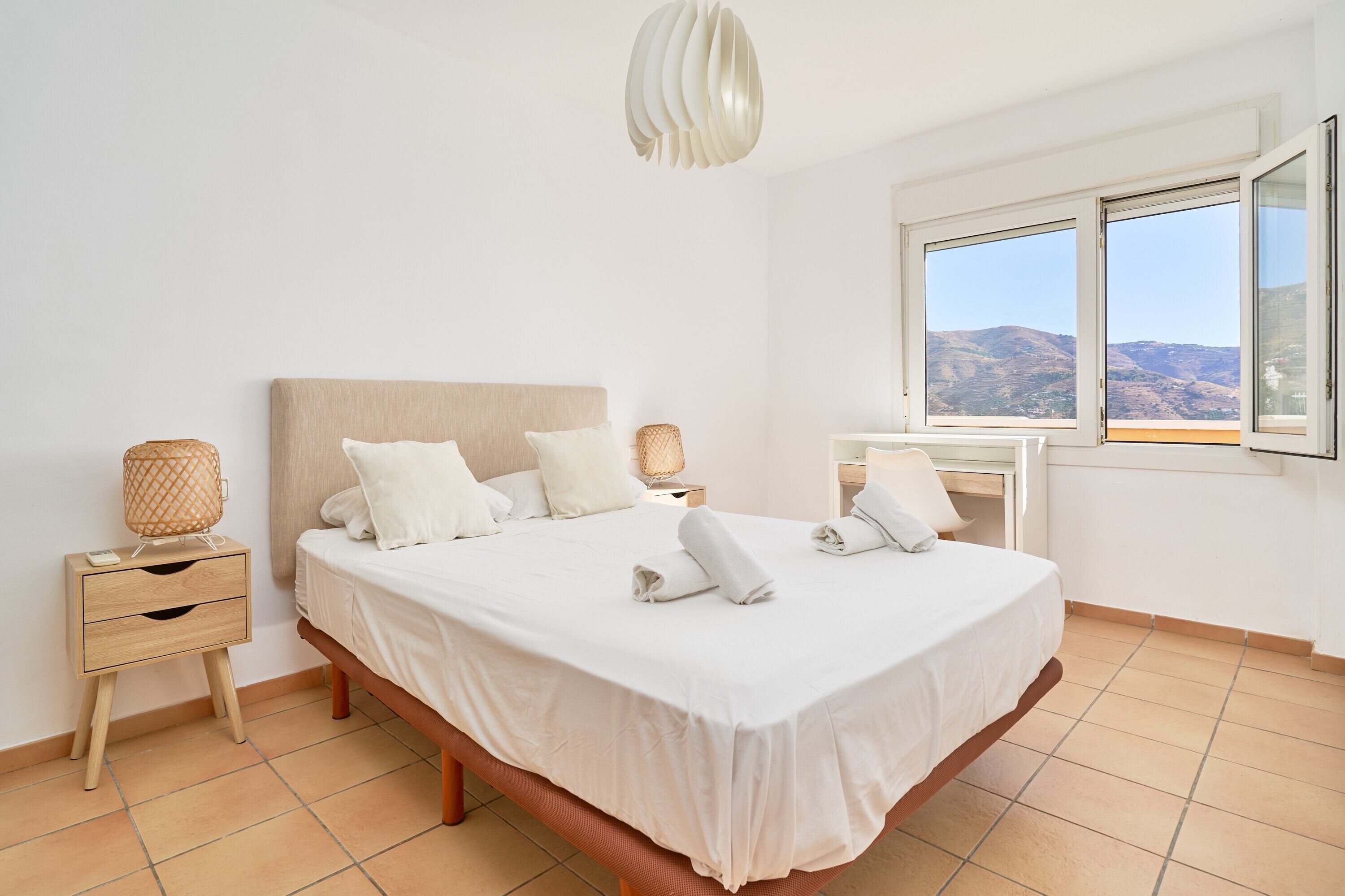 A bedroom with a bed, bedside tables, a lamp, a window with a view, and a hanging light fixture.