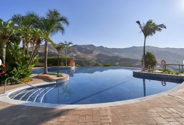 A swimming pool with clear blue water, surrounded by palm trees and a mountainous landscape in the background.