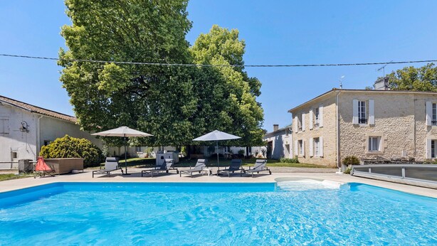 A swimming pool with lounge chairs and umbrellas, surrounded by stone buildings and trees.