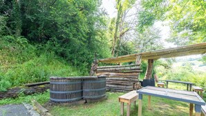 A rustic outdoor seating area with wooden benches and a table, surrounded by greenery and a log structure.
