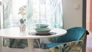 Un coin repas avec une table ronde blanche, une chaise turquoise ornée d’un motif de feuilles et un rideau à motifs tropicaux.