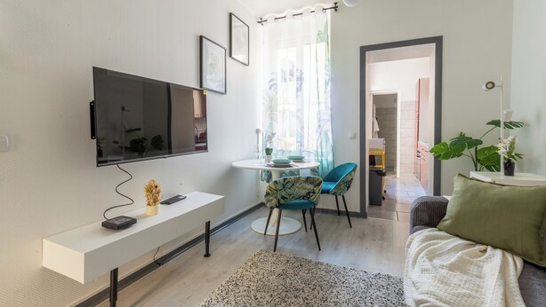 Un salon moderne avec un téléviseur à écran plat, une console blanche, des fauteuils turquoise, une grande fenêtre avec des rideaux légers et une plante verte.