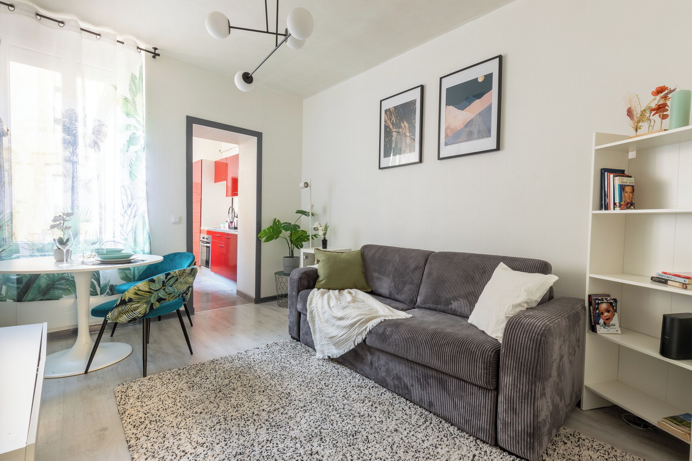 Una sala moderna con un sofá gris, una mesa de centro blanca y una silla azul. Se observa una estantería con libros y objetos decorativos, además de una planta sobre una mesita auxiliar.