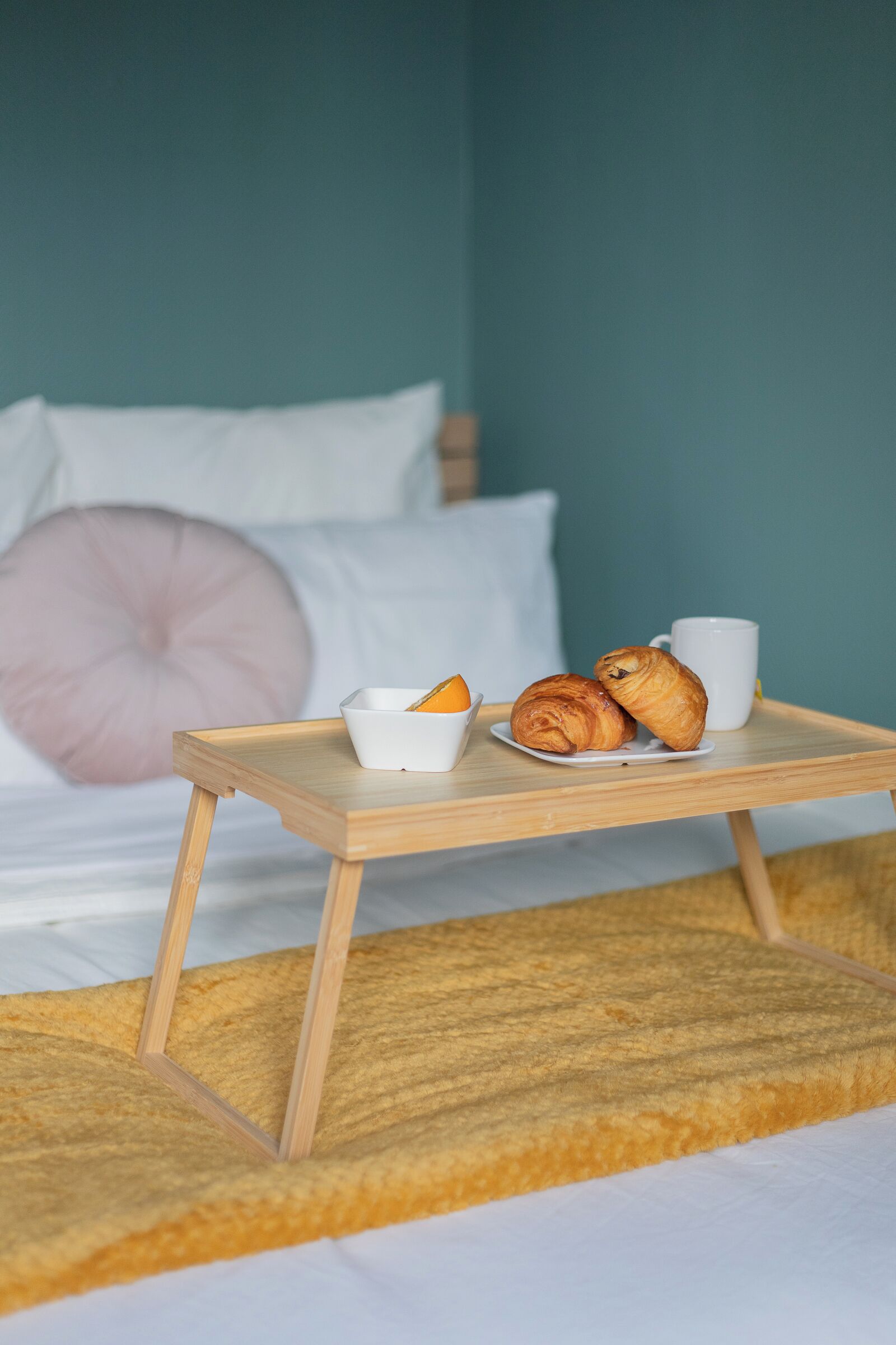 Un lit avec un oreiller rose, une table de chevet en bois sur laquelle se trouve un plateau avec des croissants et une tasse, et un bol de fruits.