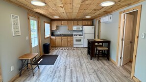 A compact kitchen with wooden cabinets, a microwave, a stove, and a refrigerator. There is a small dining table with two chairs. The floor is wooden, and there are two windows providing natural light. - Charming 2-bedroom cabin with WiFi, AC in serene Oscoda (Oscoda)