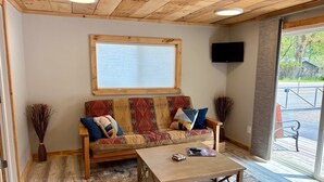 A cozy living room with a wooden ceiling, a large window with blinds, a sofa with patterned cushions, a coffee table with magazines, and a view of the outside. - Charming 2-bedroom cabin with WiFi, AC in serene Oscoda (Oscoda)